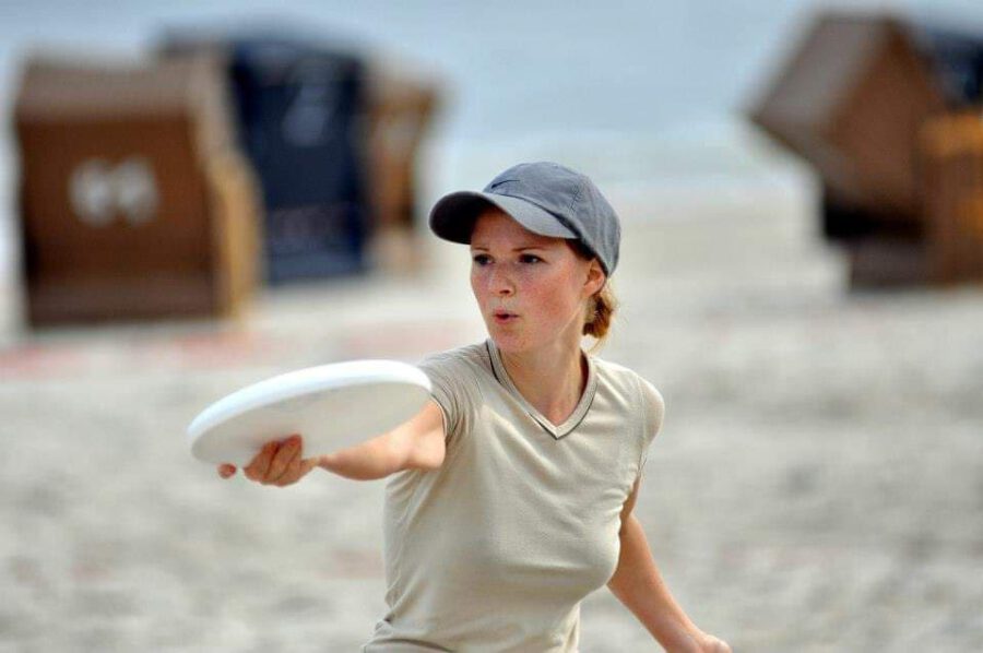 Blone, weisse Frau mit blauen Basecap setzt mit dem linken Arm zum Wurf einer weißen Frissbeescheide an. Ihr Blick ist konzentriert, ihr Mund zum ausatmen geöffnet. Im Hintergrund stehen verschwommenen Strandkörber und der Untergrund ist heller Sand.